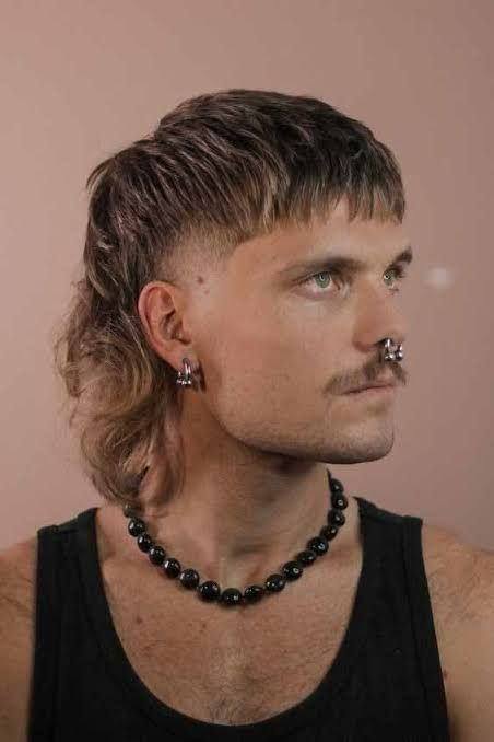 Portrait d’un homme portant une coupe mulet, un moustache fine, des piercings au nez et aux oreilles, un débardeur noir et un collier de perles noires, posé de profil sur un fond uni.