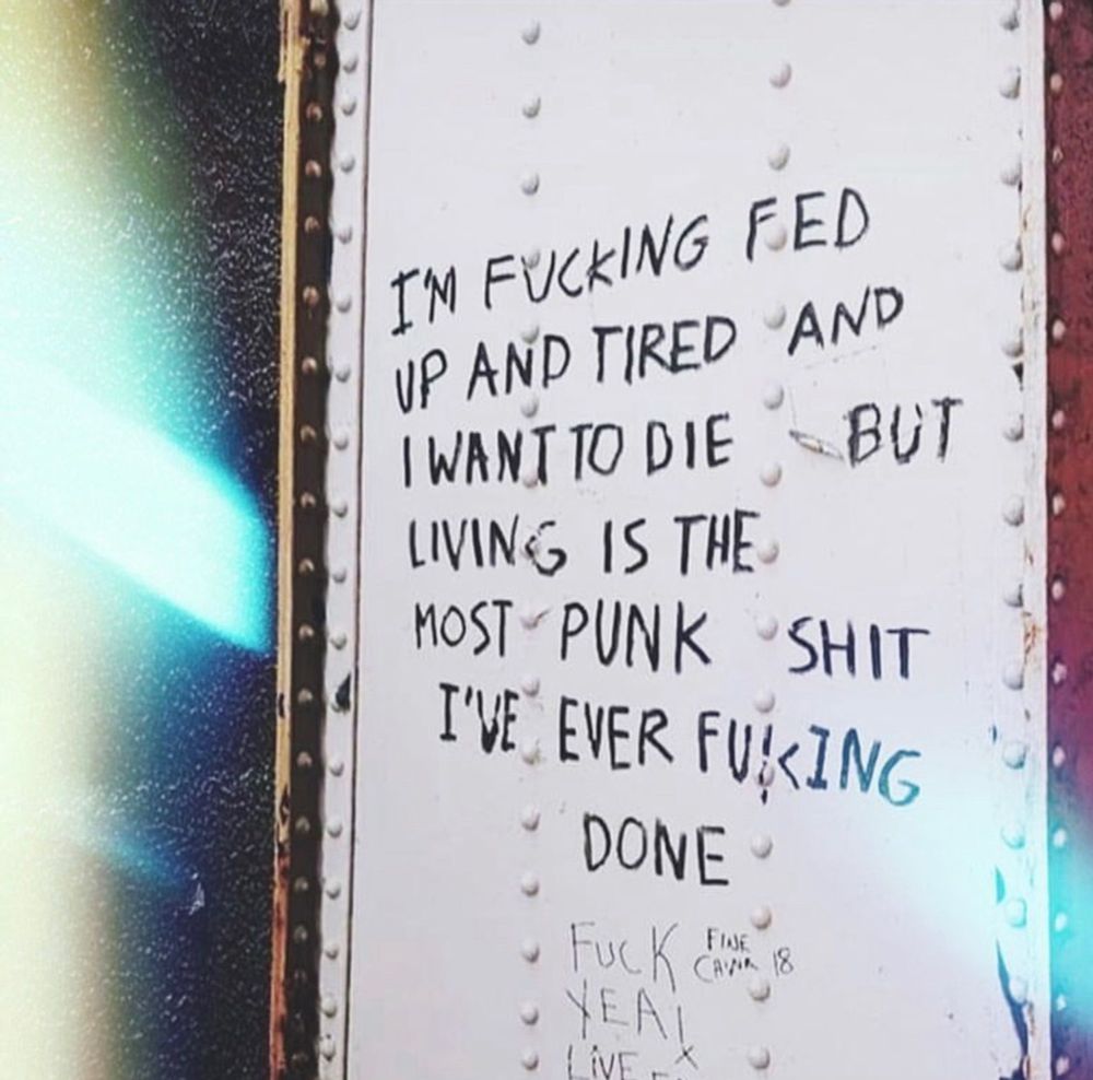 Handwritten graffiti on a wall reads
I’M FUCKING FED
UP AND TIRED AND
I WANT TO DIE  BUT
LIVING IS THE 
MOST PUNK SHIT 
I'VE EVER FUKING
DONE

Below that, someone else has scribbled
Fuck yeah!
Live