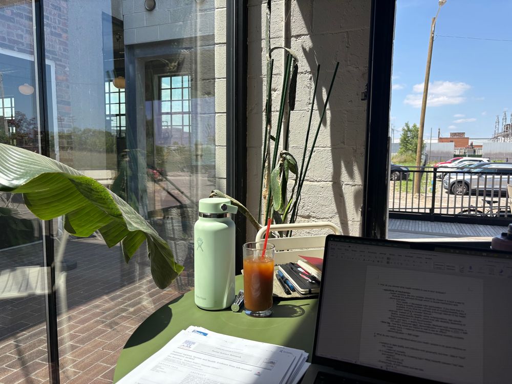 A computer, print outs of papers, plants, a green water bottle and glass of iced coffee in a sunny cafe 