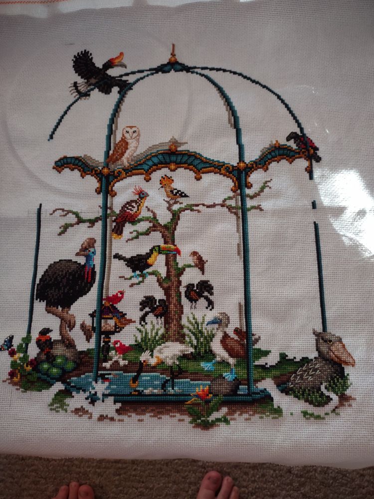Cross stitch project with aviary filled with birds and trees. Project is half done.