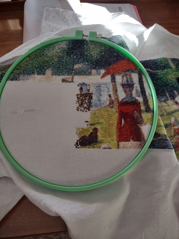 Green embroidery hoop with cross stitch. A lake a grassy area area shown with people.