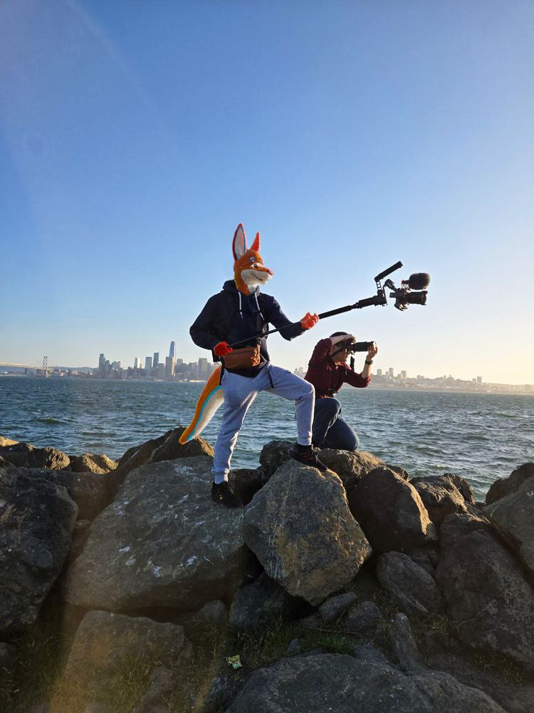 a picture of my fursona holding out a monopod with a gimbal at the end on the treasure island shoreline with san francisco in the background.