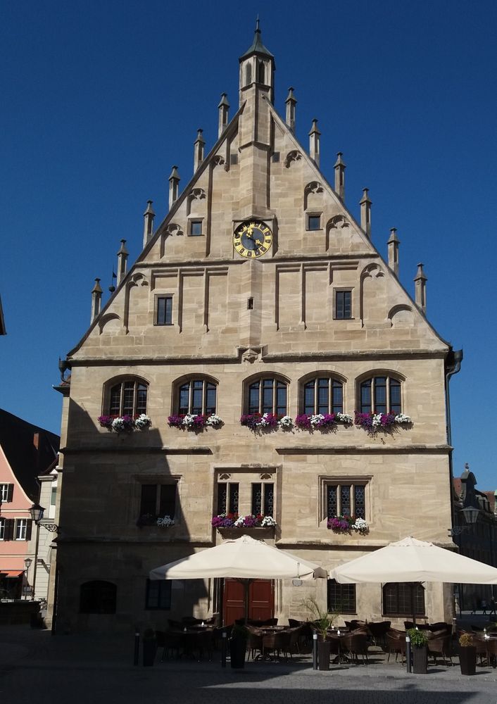 Blick auf ein weiß-beiges Backstein-Haus mit teils gebogenen Fenstern mit weißen und lila Blumen drin. Das Dach hat rechts und links schornsteinartige Aufsetzer, in der Mitte eine Uhr. Die Sonne scheint darauf, links Schatten eines gebäudes. Davor mit weißen Schirmen ein paar Stühle und Tische. Der Himmel ist leuchtend blau.