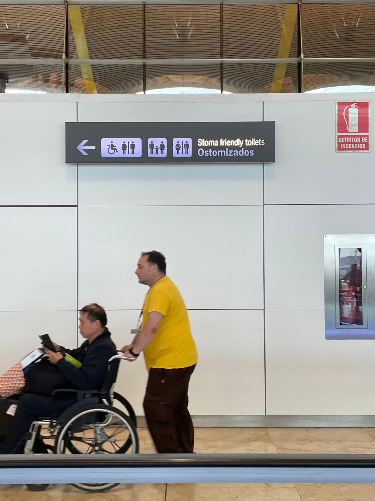 Pasillo de un aeropuerto (Barajas, Madrid). En primer plano una persona de perfil empujando una silla de ruedas que lleva a otra persona. Al fondo la típica señalización de aeropuerto indicando la dirección de los baños, añadiendo iconos de personas con ostomia y el texto “stoma friendly toilets”