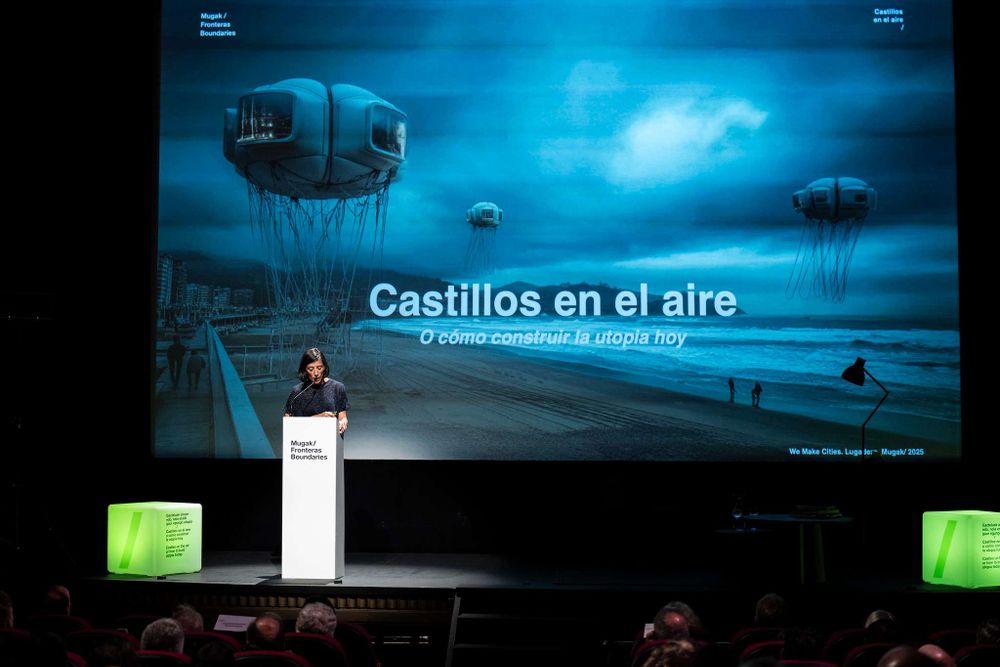 Maria Arana presentando, con una pantalla bastante grande detrás de ella, en tonos azul oscuro, con unas casas volando y el nombre "Castillos en el aire" escrito en blanco.