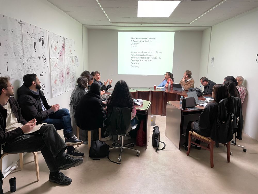 Anna Puigjaner lecturing about the kitchenless city. A medium size space, grey floor and white ceiling with a rounded table full of students, a projection of a slide in the front wall.