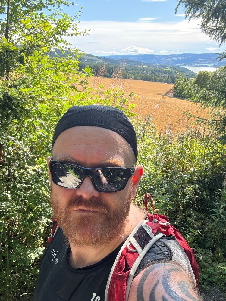 Bilde av kjekkebass på tur. Mann i sin beste alder med solbriller og ryggsekk. I bakgrunnen kan vi se et solfylt Lillehammer-landskap.