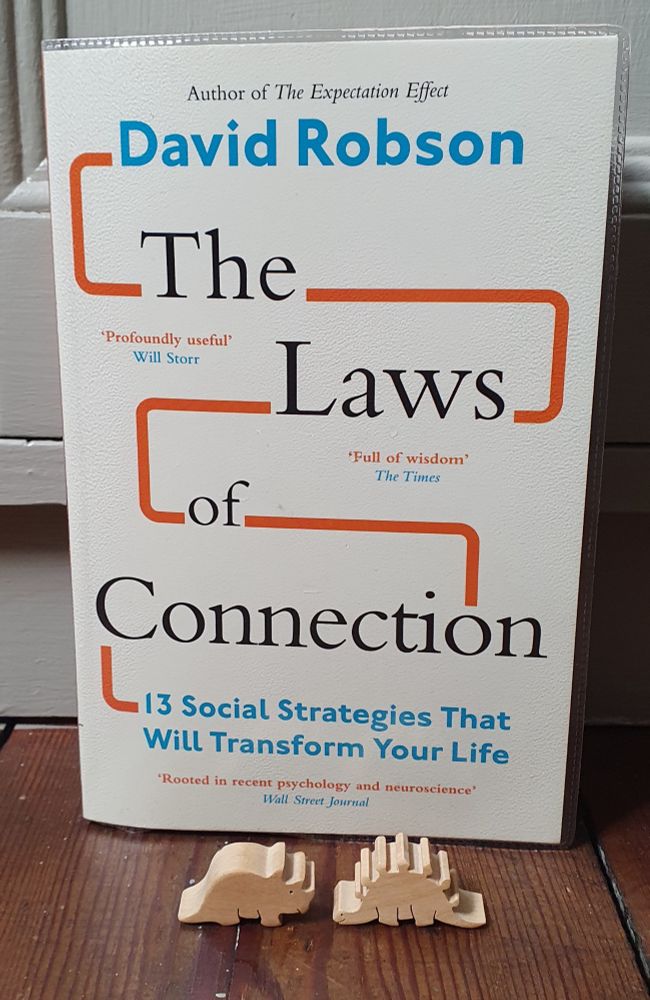 Paperback copy of The Laws of Connection: 13 Social Strategies That Will Transform Your Life by David Robson. In front of the book are two small dinosaurs about to touch noses.