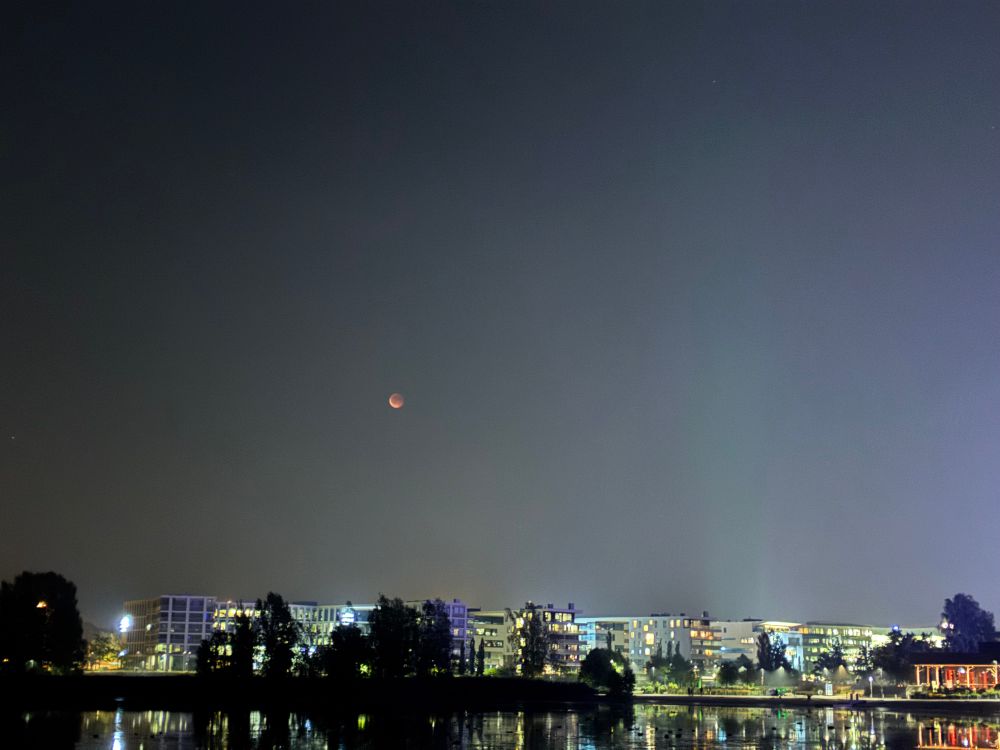 A nighttime shot of a city skyline. The Moon appears low in the sky and is in full eclipse, all red, brighter at the bottom left and darker towards the upper right.