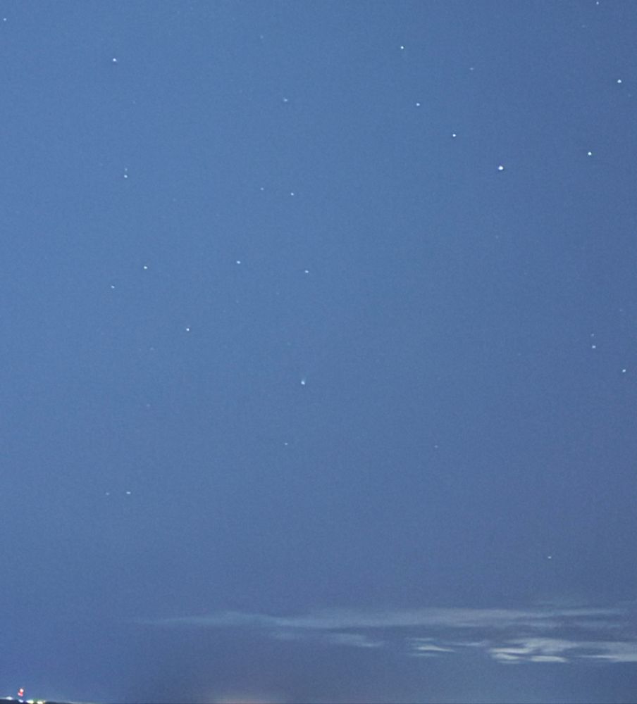 A blue-tinted image of the night sky, filled with stars. Near centre, there is a small smudge that is a comet.