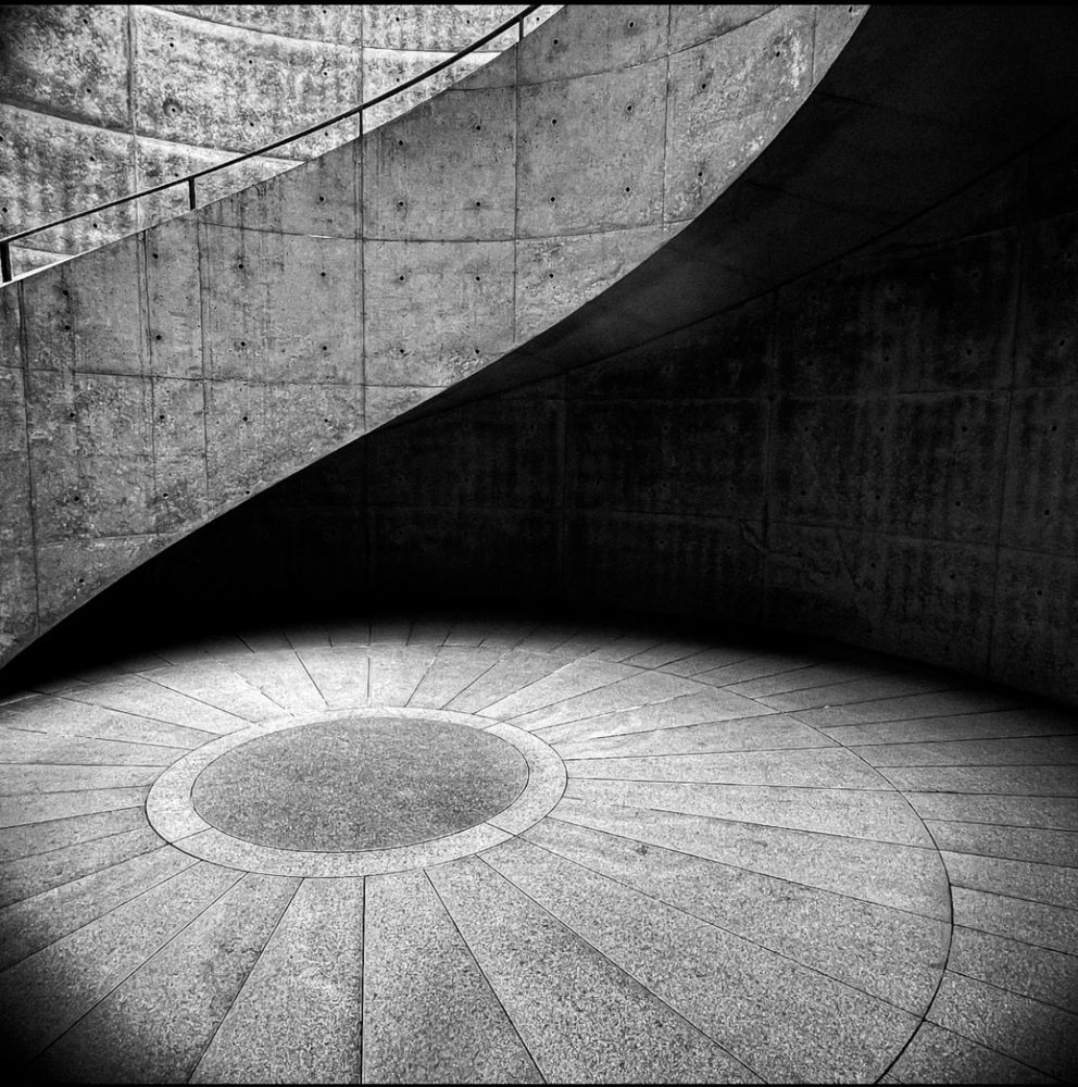 black and white photo of a staircase following a curved wall. there is a circular pattern on the floor that seems to connect to the spiral of the stairs 