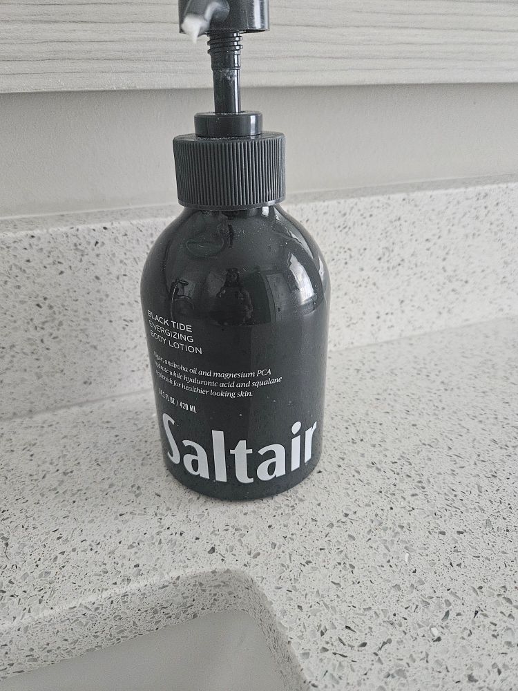 A dark gray lotion bottle, sitting on a marble countertop. There is large text at the bottom of the bottle that reads "Saltair".