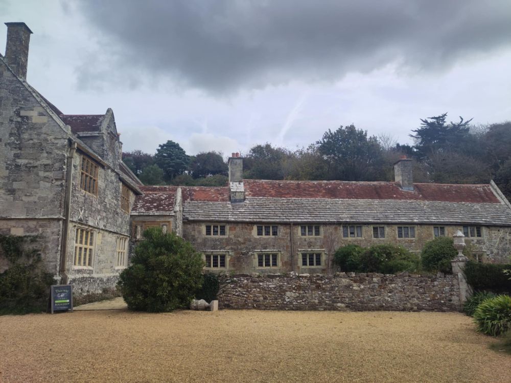Photo of Mottistone Manor house in the Isle of Wight. It was first mentioned in the Domesday Book of 1086, but most of the building dates from the 15th and 16th centuries. The building has two wings, south-east and north-west, both seen here, with two floors and a low roof. Above the house is a sky moody as a teenager.
