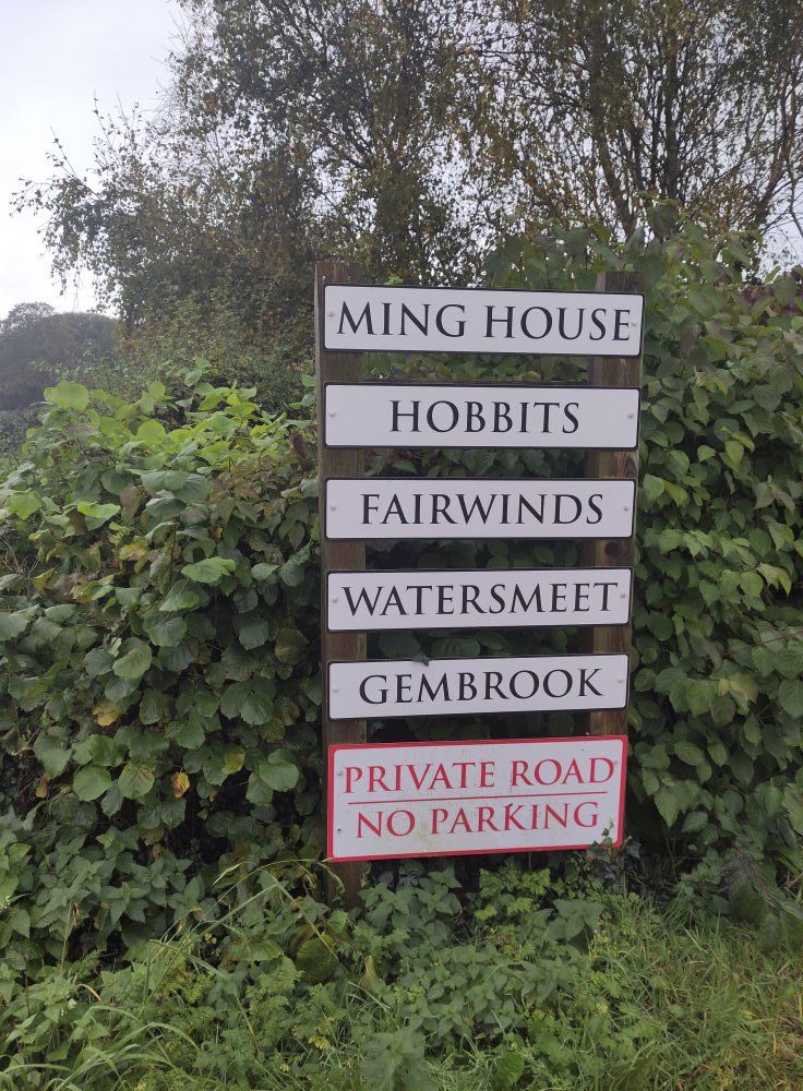 Photo: a street sign plus house name signs, arranged perpendicularly, in Brighstone, near Newport, Isle of Wight.
The bottom sign, red lettering with a red border, says: private road, no parking.
The other signs, reading from top downwards, are:
Ming House,
Hobbits,
Fairwinds,
Watersmeet,
Gembrook.
