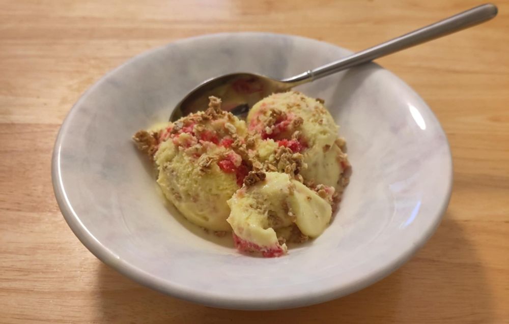 A bowl containing three scoops of icecream. The icecream is yellow with a red ripple through it and is scattered with toasted oats. There is also a spoon sticking out of the bowl.