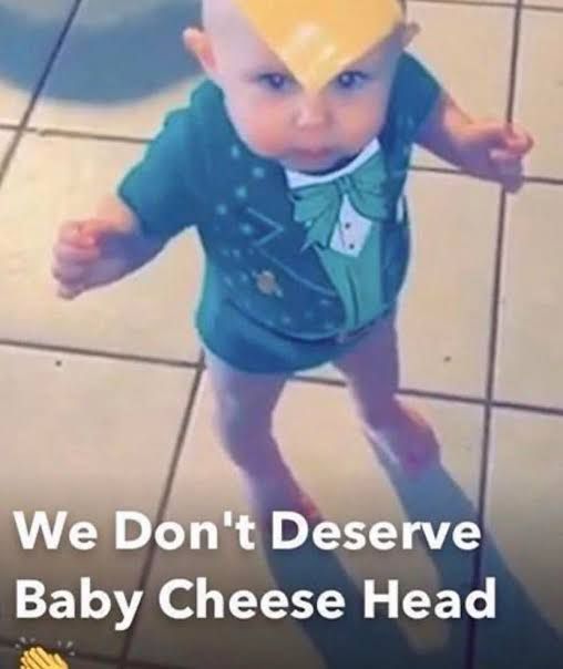 A toddler standing with a slice of cheese on their head. The text reads ‘We Don’t Deserve Baby Cheese Head 👏’