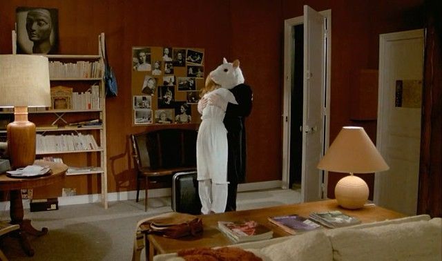 Photogramme du film d’Alain Resnais Mon oncle d’Amérique. Dans un salon années 80 aux teintes orangées, une femme de dos, en robe blanche, est étreinte par un personnage vêtu de noir à la tête de souris blanche géante.