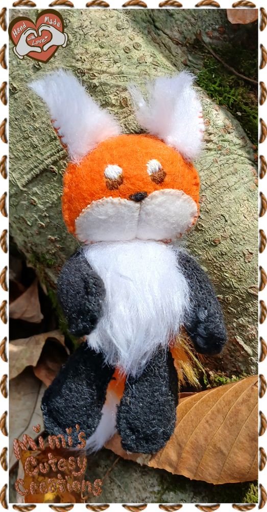 A small orange fox plushies from a front view. It has a white muzzle, fluffy white inner ears, white neck fluff, and black arms and legs. it is shown in front of tree bark and dried brown leaves.