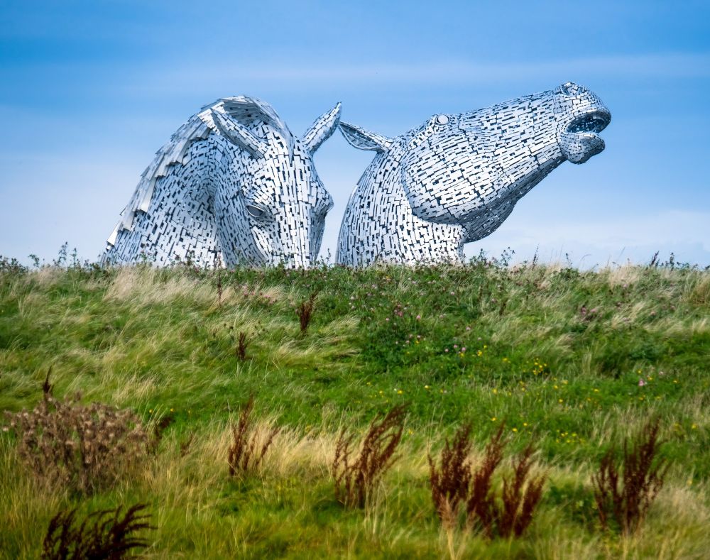 A large metal sculpture of two horse heads that appear to be coming out of the ground with a green grassy hill in the foreground.