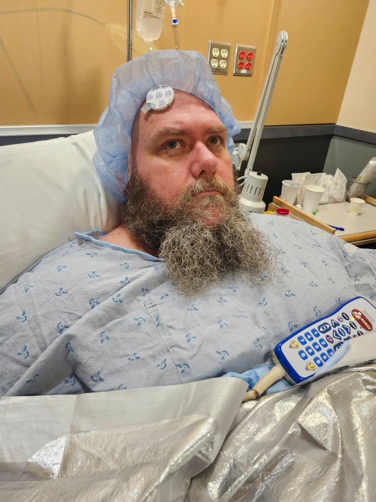 A bearded man in a hospital gown in a hospital bed waiting for surgery 