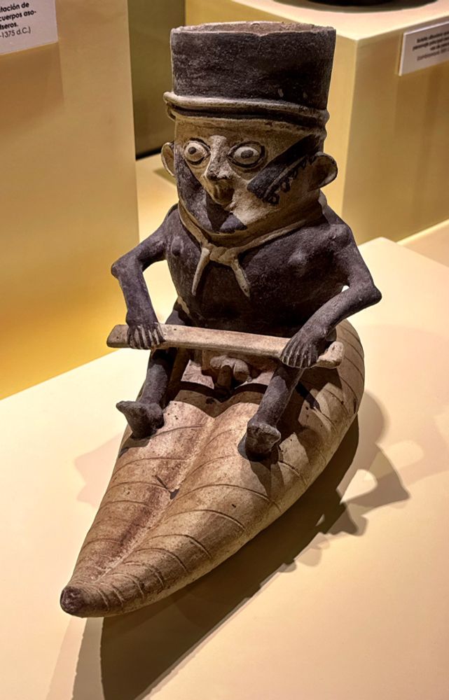 A naked man with the fixed grin of someone posing for a portrait with his genitals slapped on an unfinished reed boat.