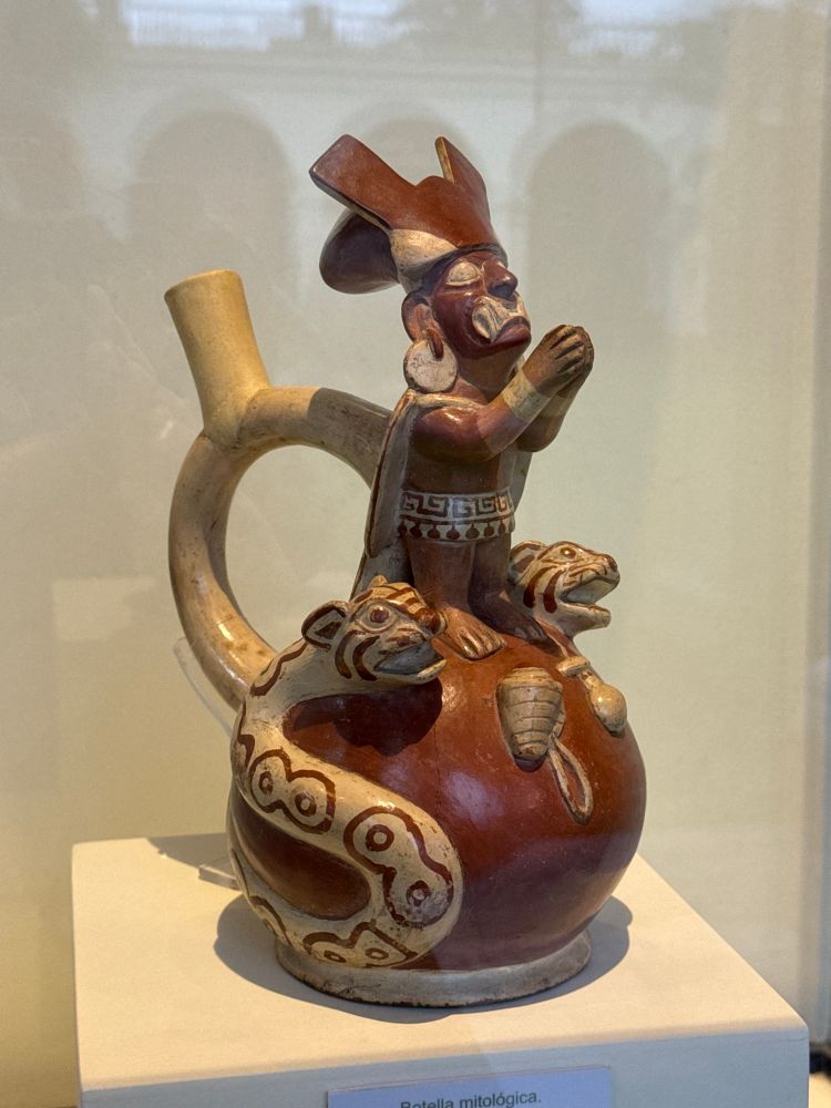 A pot showing a figure with a large headdress and clasped hands. Two snakes with snarling feline heads wrap around the base of the bottle.