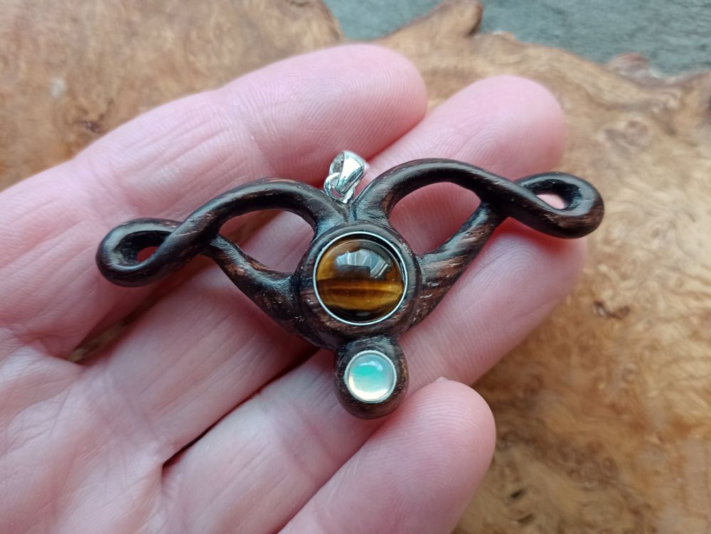 A wooden piece of jewellery, consisting of two swirling arms, with a round tiger eye gemstone in the centre, and a smaller round opal gemstone underneath. On the fingers of a hand, with a wood background.