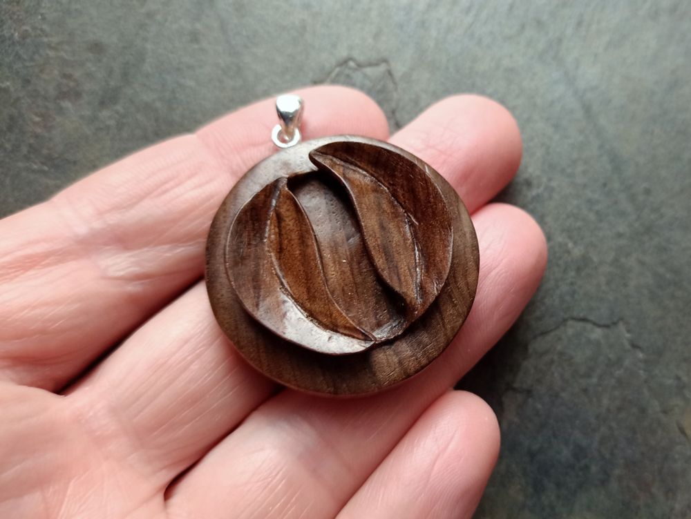 A round wooden pendant, carved from a rich brown walnut, of two leaves carved in relief, circling each other , so the tip of each leaf ends at the stalk of the other. Resting on the four fingers of a hand, on a grey slate background.