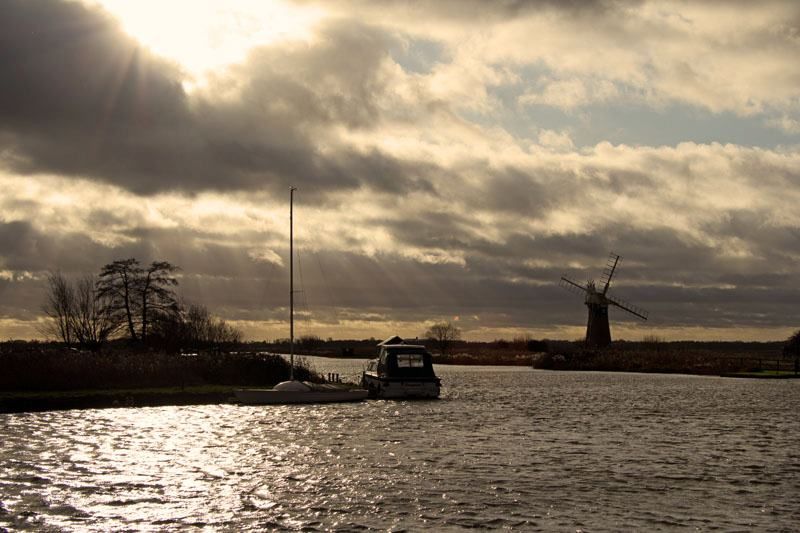 A view from beyond the bend of a river in the Norfolk Broads. On the inside of the bend, to the left, are moored a small cabin cruiser and a sailing boat. A wind pump or mill can be seen by an inlet, just beyond the bend and to the right. Dark clouds subdue the overall lighting, however the sun breaks through from above. Photograph by Magusian.