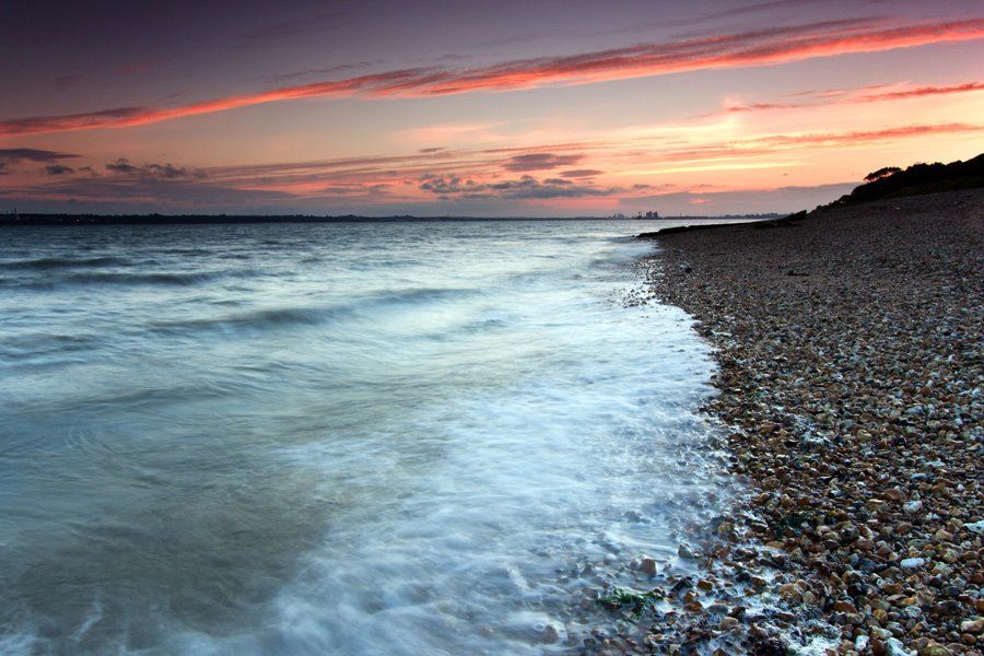 The sun has set and the scattered clouds above reflect deep reds and oranges, whilst floating in the darkening sky… thus reminding us that the sun continues to radiate its light, despite being obscured by the horizon and the distant line of clouds.

The view is from a sloping pebble beach and extends along the estuary towards the distant town of Southampton. A timed exposure is used to slow down the moment, and to soften and smooth the appearance of the waves as they break along the beach. A long-exposure photograph by Magusian.