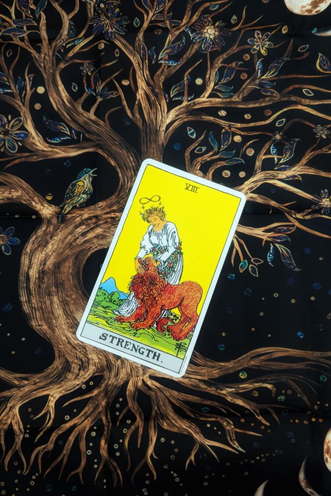 The image shows the Strength card from the Original Rider-Waite Tarot Deck, illustrated by Pamela Colman Smith, and photographed on a reading cloth that features the World Tree, complete with a singing bird.