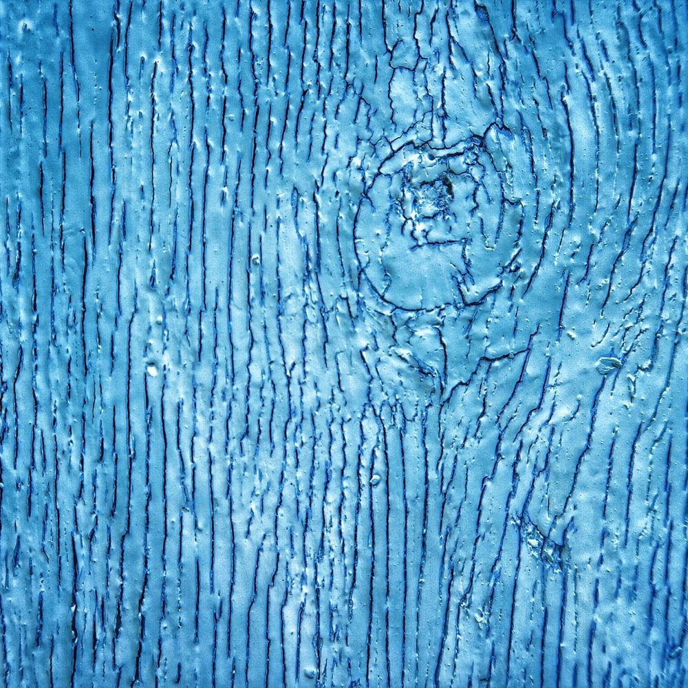 A close-up photograph of a section of wood, which forms a part of an old shed. The blue paint is faded and cracked, with textures from the grain and knot enhanced by the oblique sunlight. Taken by Magusian.