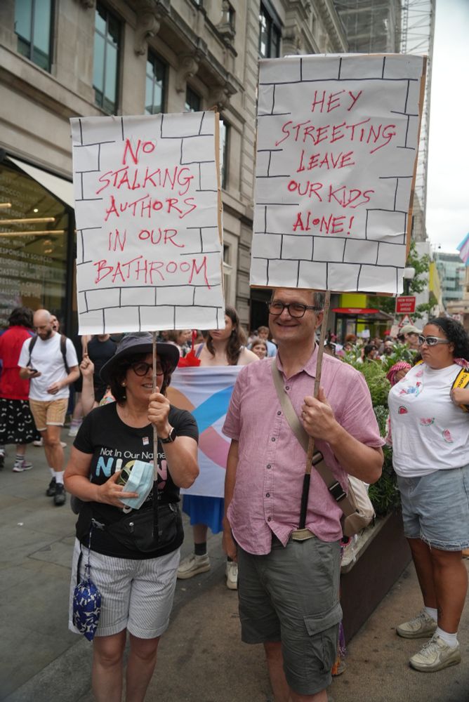 The same people with the same protest signs but this time the left sign has been flipped to show what's written on the back. It's once again a hand drawn brick wall texture on which text is written. The text says "No stalking authors in our bathroom"