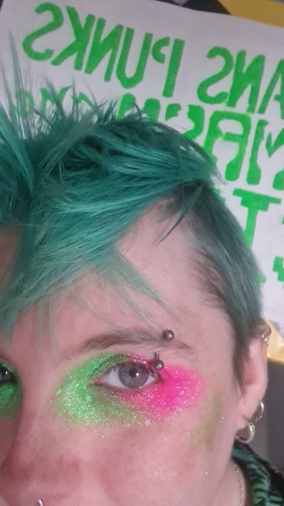 Close up shot of Flint from WhatTheTrans, half his face is out of frame, only really showing the green hair on top and one eye with green and pink glitter on it. In the background is a sign reading "Trans Punks Smash the Cistem"