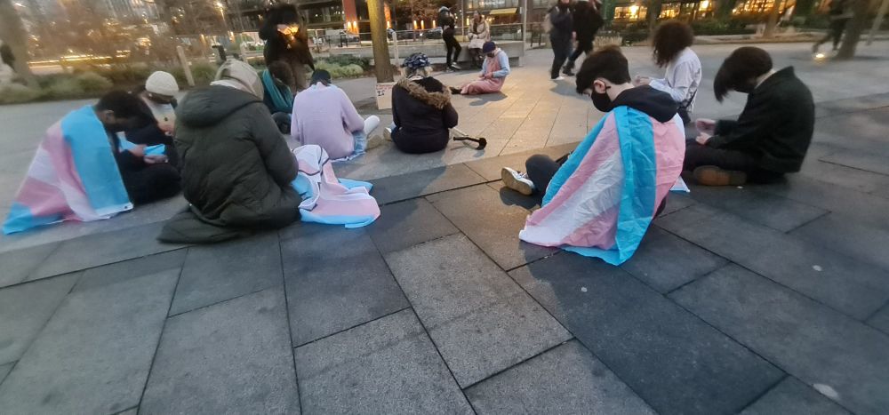 Protesters sat down outside the embassy with trans flags making bracelets 