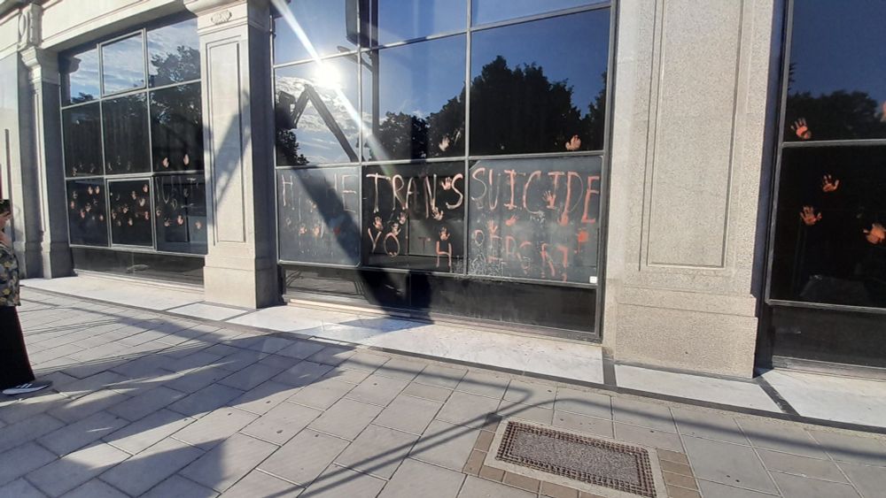 The front of NHS England, covered in bloody handprints. A message written in red says, "Release the trans youth suicide report.