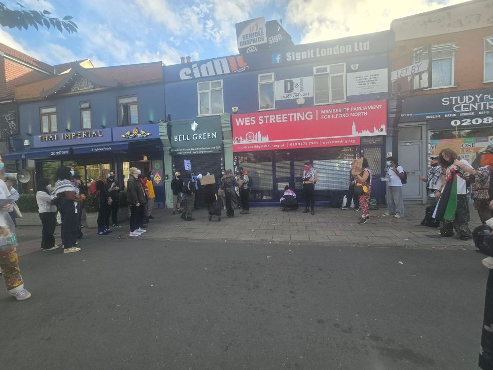 A group of people gathering outside of wes Streeting's office. A building with a red sign on the top saying "Wes streeting" on it. 