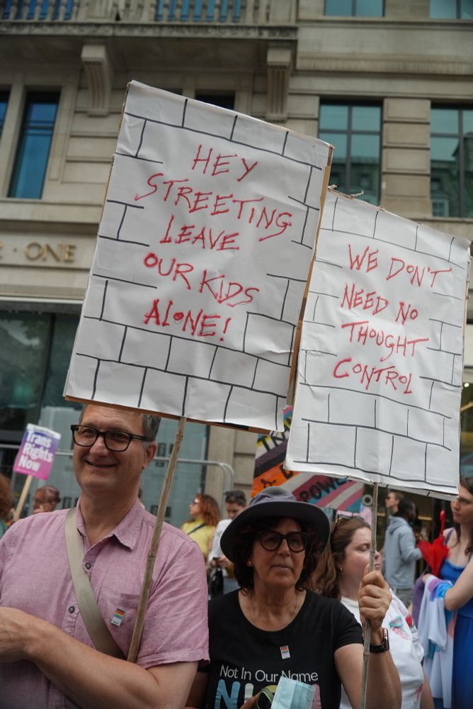 Two people holding matching protest signs with text over a hand drawn brick wall texture. The left sign says "Hey Streeting Leave Our Kids Alone!" The right one says "We don't need no thought control!