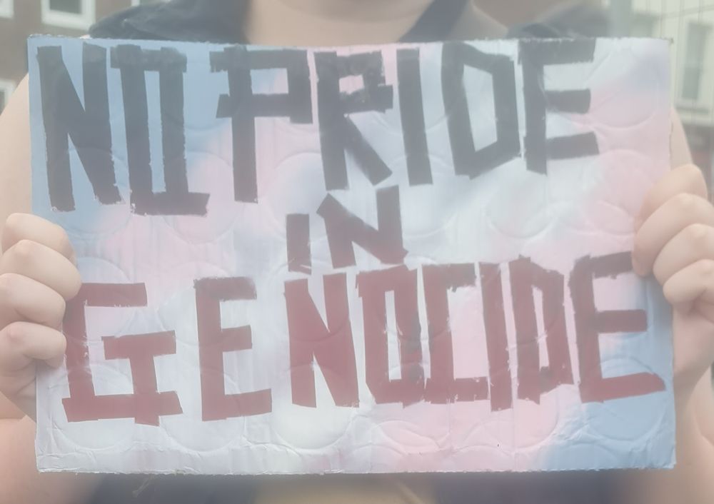 A white person holding a sign saying "no pride in genocide" with a trans flag background 