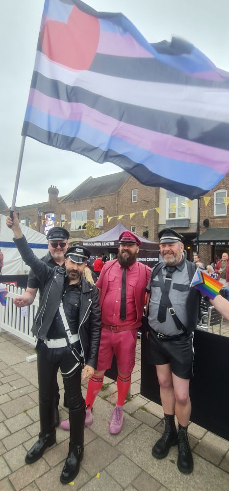 Four leather daddies, dressed in the classic style with black leather jackets, ties and hats, one in all pink. One member of the group is waving a leather daddy flag, which is usually a heart in the top left corner, with blue and black stripes as the background, but this flag has black, blue, pink and white stripes, in the shades used for trans pride flags