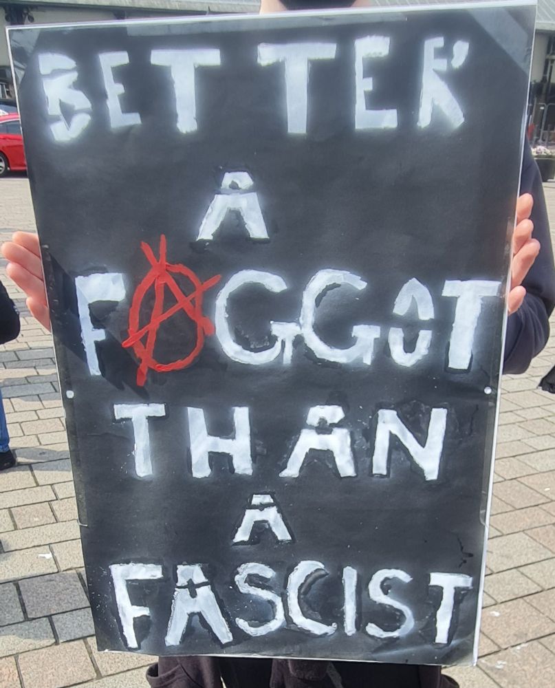 A person wholding a placard, you cannot see their face, only the tips of their fingers, they are holding a large black sign with white lettering saying "Better a Faggot than a Fascist" the letter A in faggot is a red anarchy symbol 