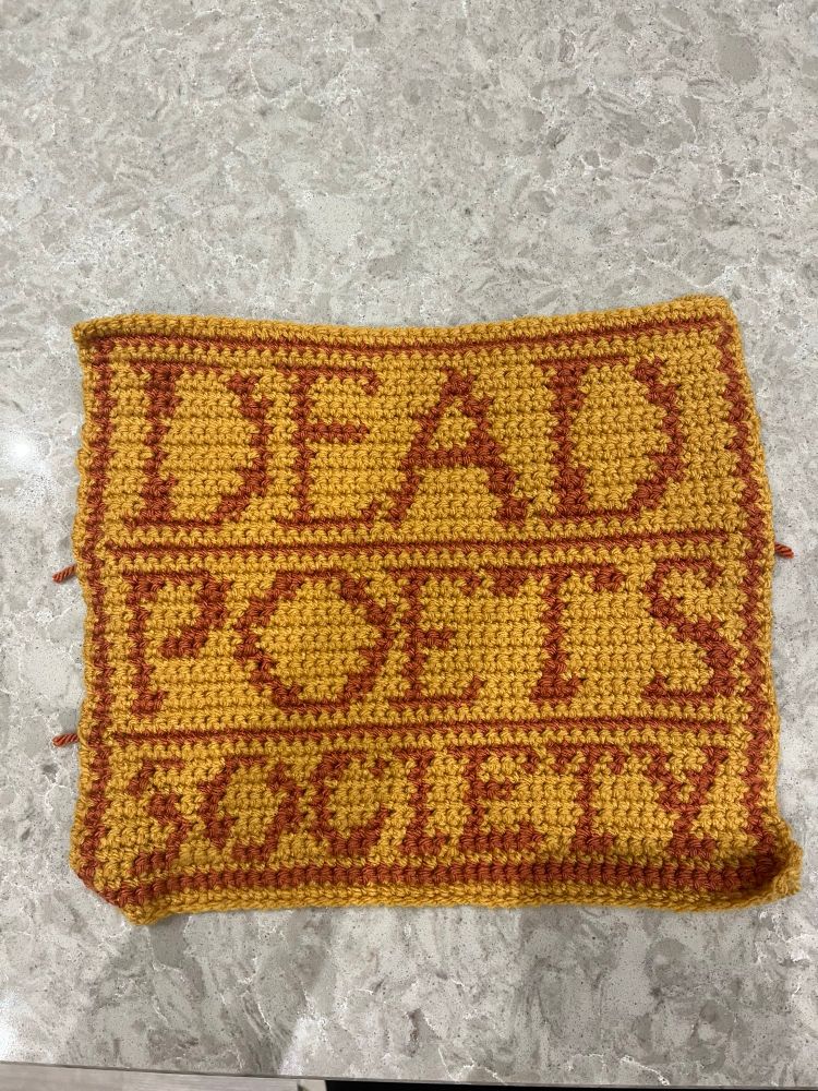 A crocheted blanket square containing the title Dead Poets Society from the film’s poster