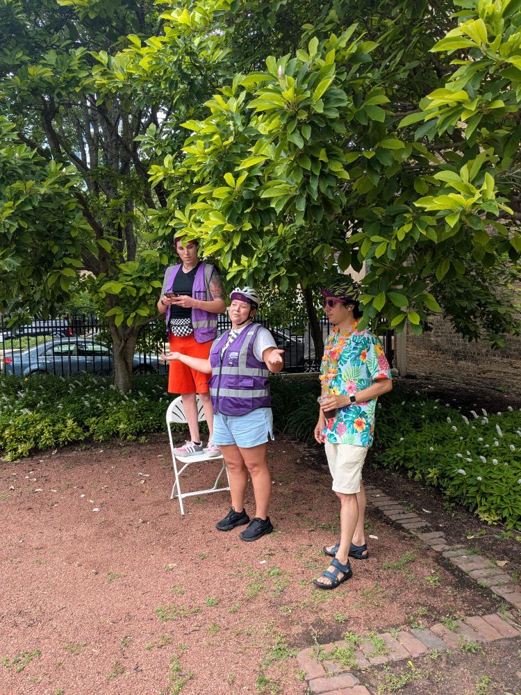 Three ride organizers under a tree giving a safety talk before the ride. Two wearing purple vests and the third in a Hawaiian shirt, one standing on a chair 