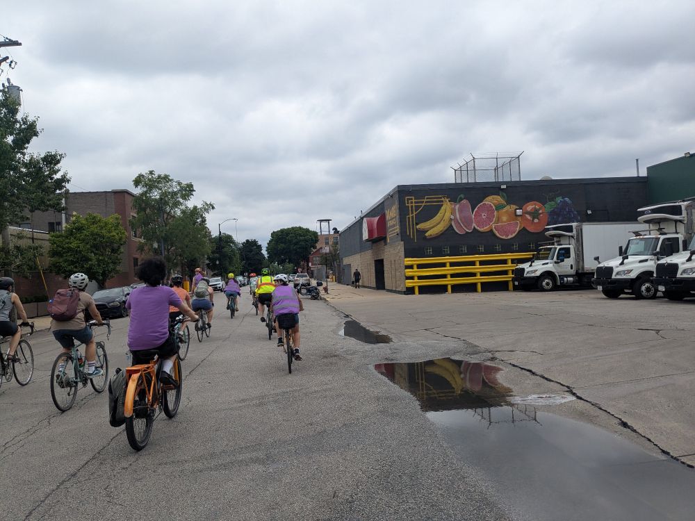 A group of cyclists riding through an industrial area with a mural of fruit on a building nearby 