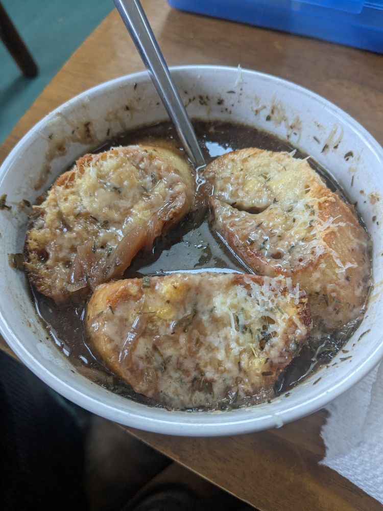 A bowl of French onion soup, a dark brown soup with three pieces of cheesy toast on top in a white ceramic bowl on a brown table 