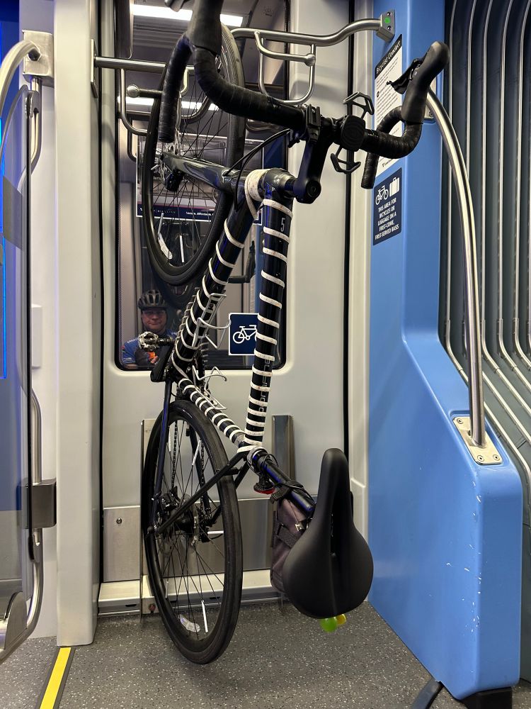 Picture of a road bike hanging on a rack in a light rail car with it's riderreflected on the window just behind the bike