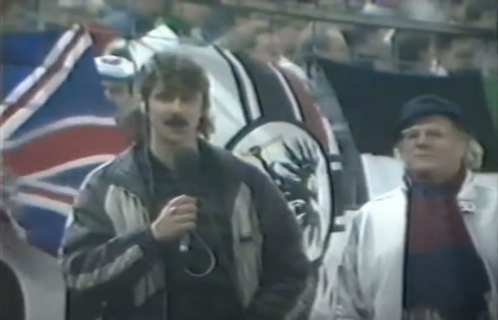 Reporter mit Mikrofon und Interviewpartner im Fußballstadion, im Hintergrund hängen die Reichskriegsflagge und der Union Jack