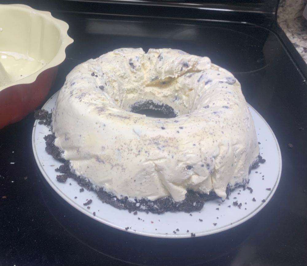 a picture of an oreo ice cream bundt cake that i made. the ice cream cake itself is on a plate on top of my stove and you can see the bundt cake mold in the background. the shape of the cake is unfortunately not the same of the bundt mold but now i know not to use parchment paper for next time. 
