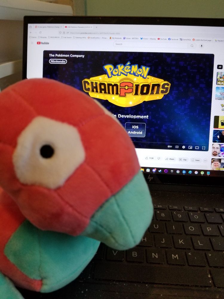 A Porygon plush in front of a laptop screen showing the announcement of Pokemon Champions in development for Switch and smartphones.