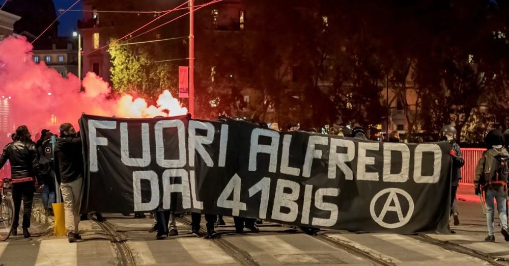 Eine Gruppe Demonstrierender blockiert eine nächtliche Strassenkreuzung und hält ein großes schwarzes Banner mit der weissen Aufschrift 
"FUORI ALFREDO
DAL 41BIS“
und einem Anarchie-Symbol. Hinter ihnen leuchtet dichter roter Rauch aus Bengalos.
